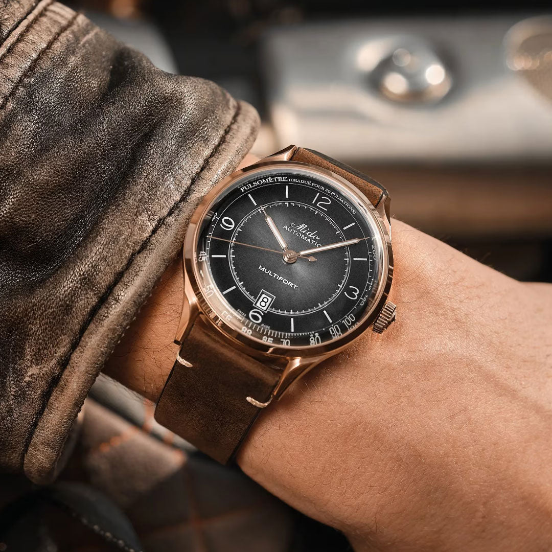 A close-up of a wrist wearing the MIDO Multifort Patrimony Automatic 40mm Watch with a brown leather strap, paired with a brown leather jacket; part of a car interior appears in the background.