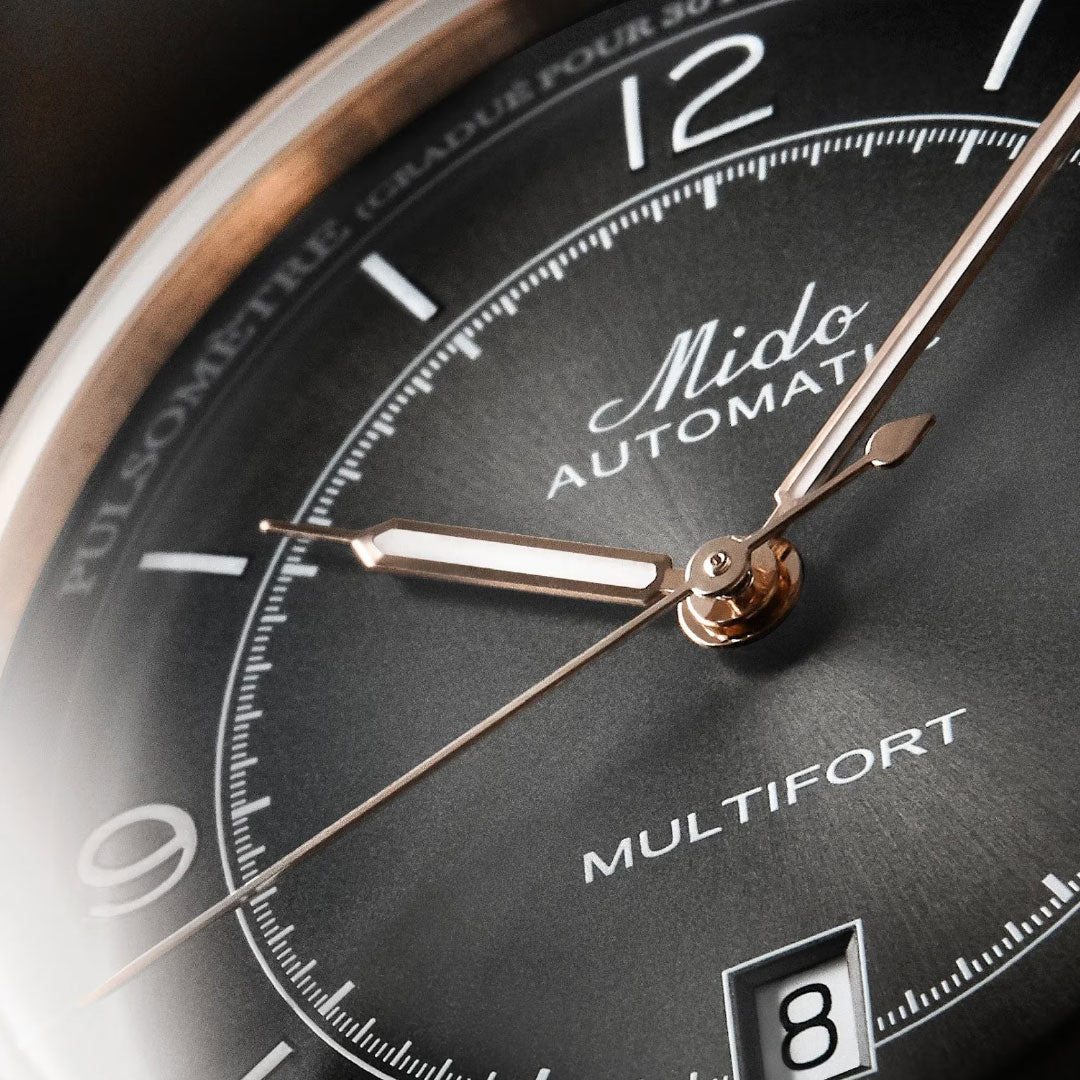 Close-up of the MIDO Multifort Patrimony Automatic 40mm Watch, showcasing gold hands, a black dial with white markings, a date window at 6 o’clock, and the detailed textures of this MIDO timepiece powered by Caliber 80 automatic movement.