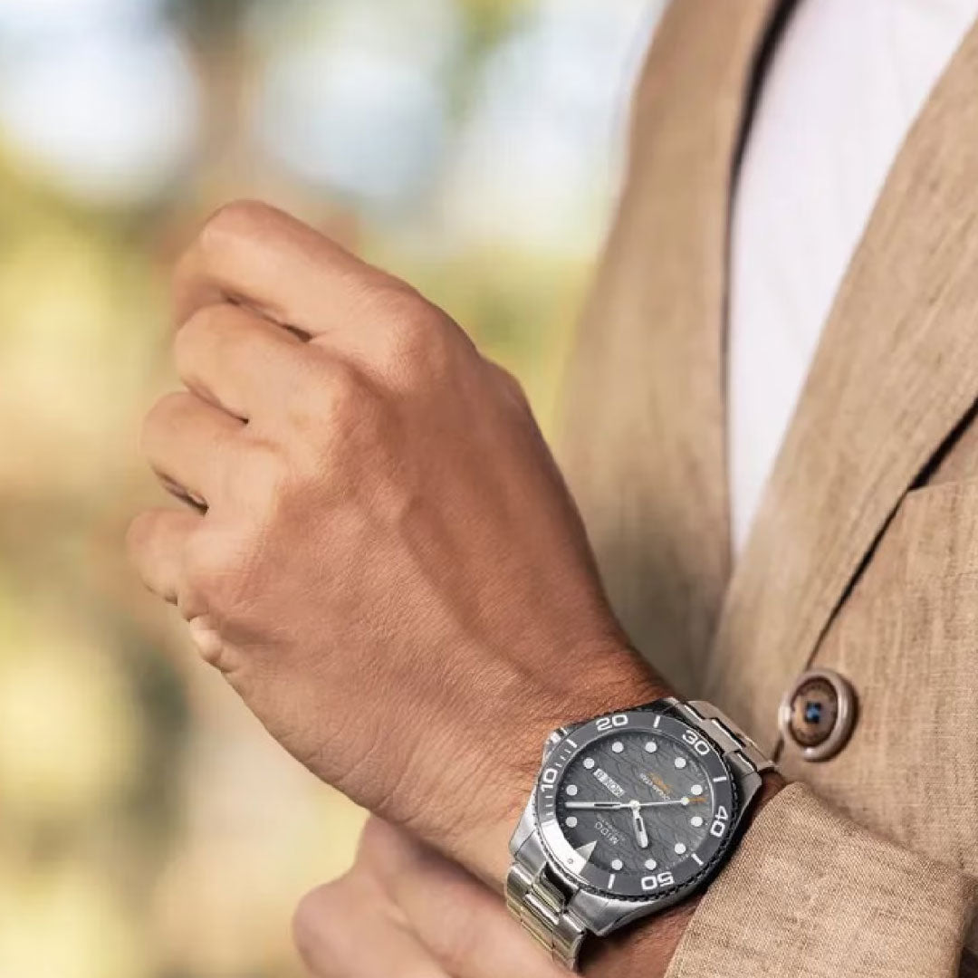 A person in a beige suit adjusts their sleeve, revealing a MIDO Ocean Star 200C Automatic 42.5mm Watch with a ceramic bezel, black dial, and white markers by MIDO, set against a softly blurred green and yellow background.