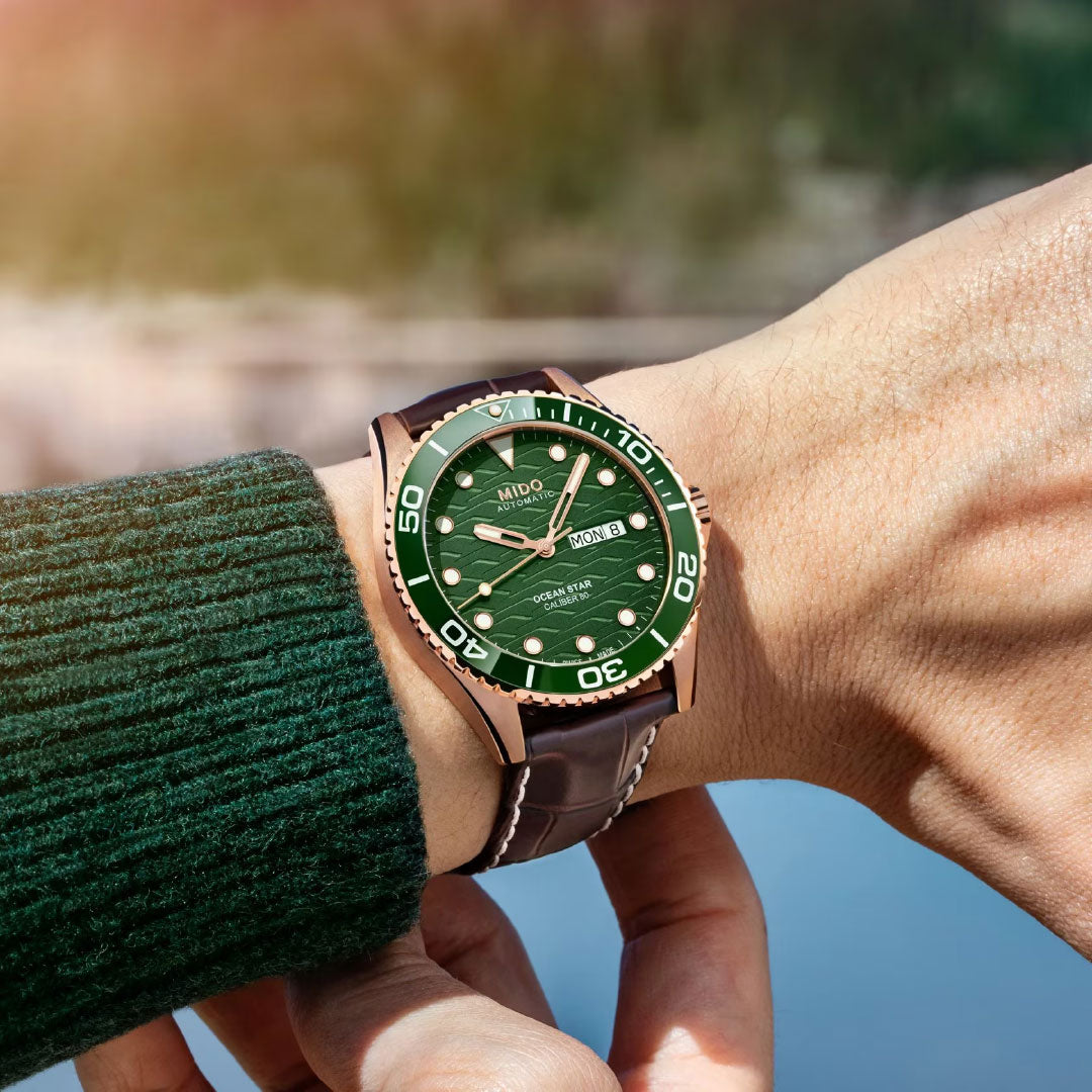 A person wears a MIDO Ocean Star 200C Automatic 42.5mm watch with a green dial and brown leather strap, standing near water with their arm outstretched against a lush forest backdrop.