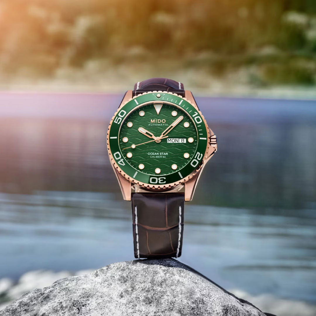 A MIDO Ocean Star 200C Automatic 42.5mm watch with a green dial, ceramic bezel, and brown leather strap is set on a rock, with a blurred body of water and trees in the background.