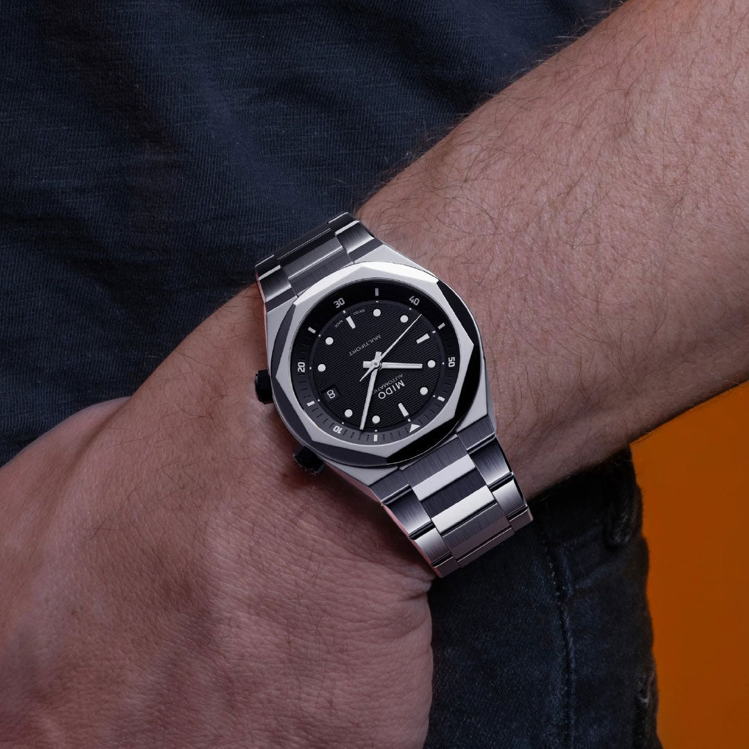 A person in a dark short-sleeve shirt stands against an orange background, wearing the MIDO Multifort 8 Two Crowns Automatic 40mm watch with a silver case, black dial, and silver hour markers.