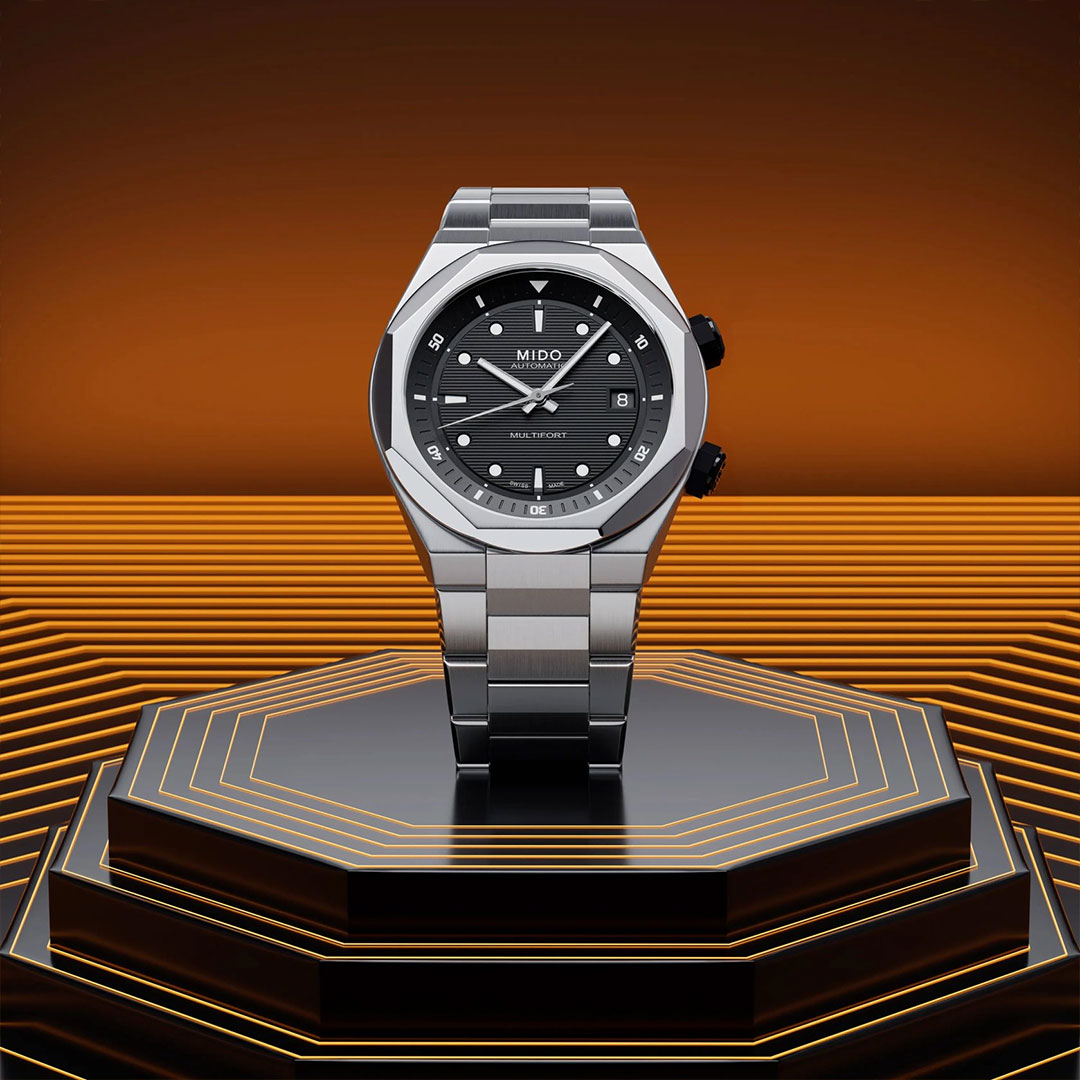 A MIDO Multifort 8 Two Crowns Automatic 40mm watch with a black dial is displayed upright on a hexagonal black, orange-lit platform against a gradient orange background featuring geometric layered patterns.