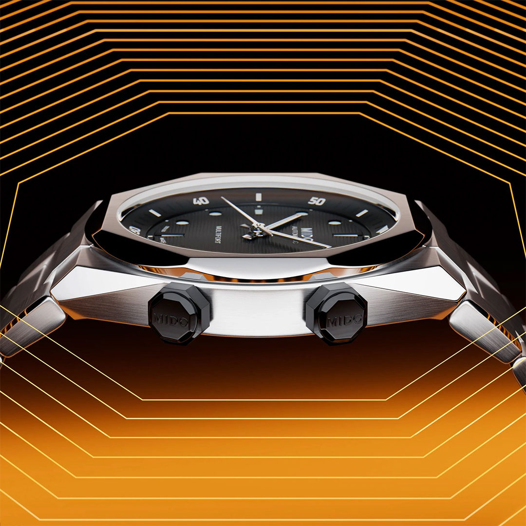 A close-up of the MIDO Multifort 8 Two Crowns Automatic 40mm Watch, featuring a black dial and metallic case, is set against orange and yellow geometric hexagonal patterns.