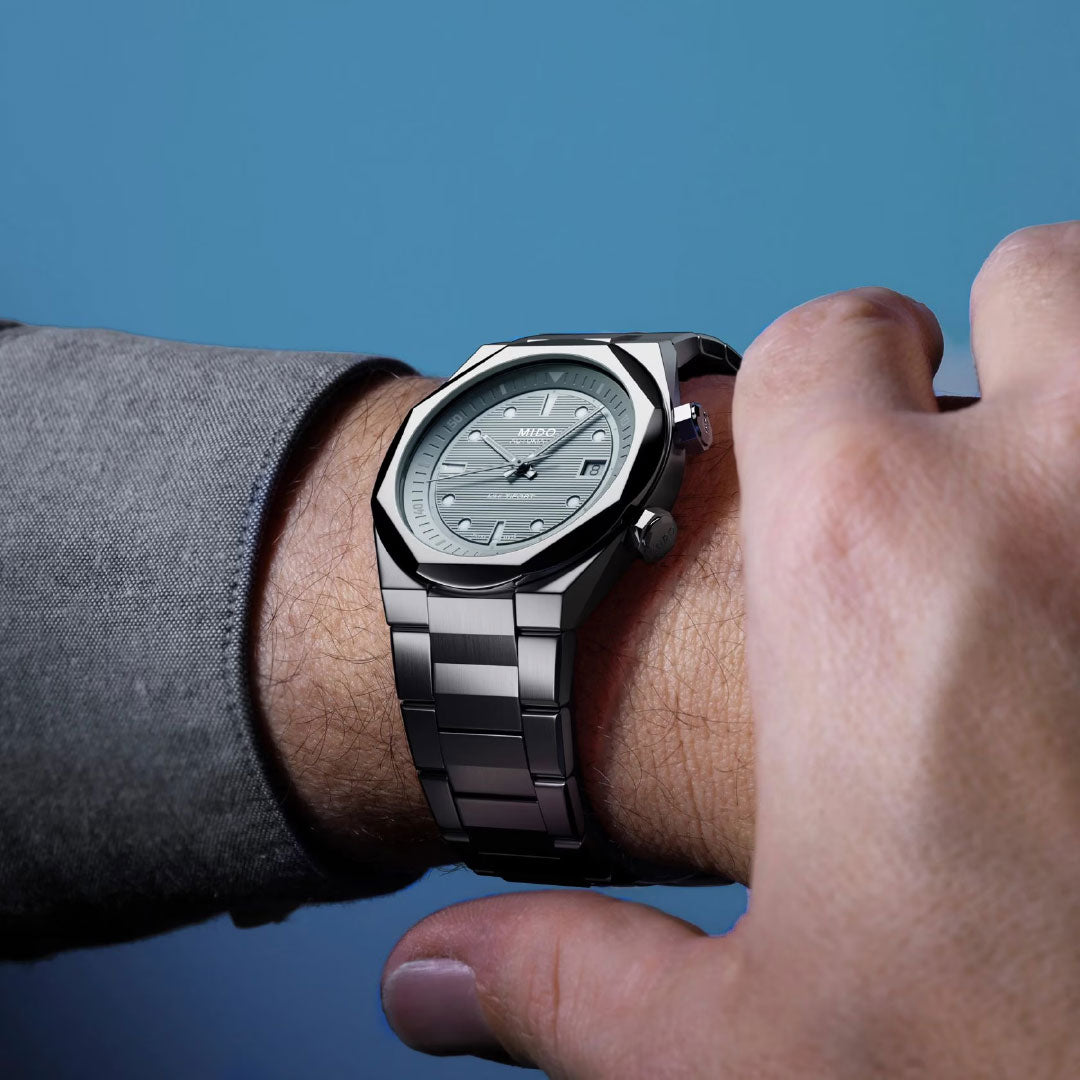 A close-up shows someone adjusting their MIDO Multifort 8 Two Crowns Automatic 40mm Watch with a metal band and gray dial on their left wrist, set against a blue background.