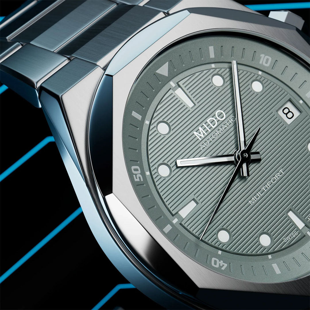 Close-up of the MIDO Multifort 8 Two Crowns Automatic 40mm watch, featuring a textured light green dial, silver hour markers, and a metal bracelet by MIDO, set against diagonal blue lines.