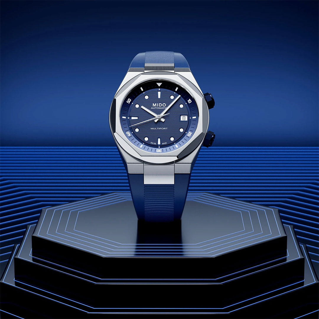The MIDO Multifort 8 Two Crowns Automatic 40mm watch, featuring a blue and silver design with a textured blue dial, is displayed on a futuristic octagonal platform against a geometric blue background.