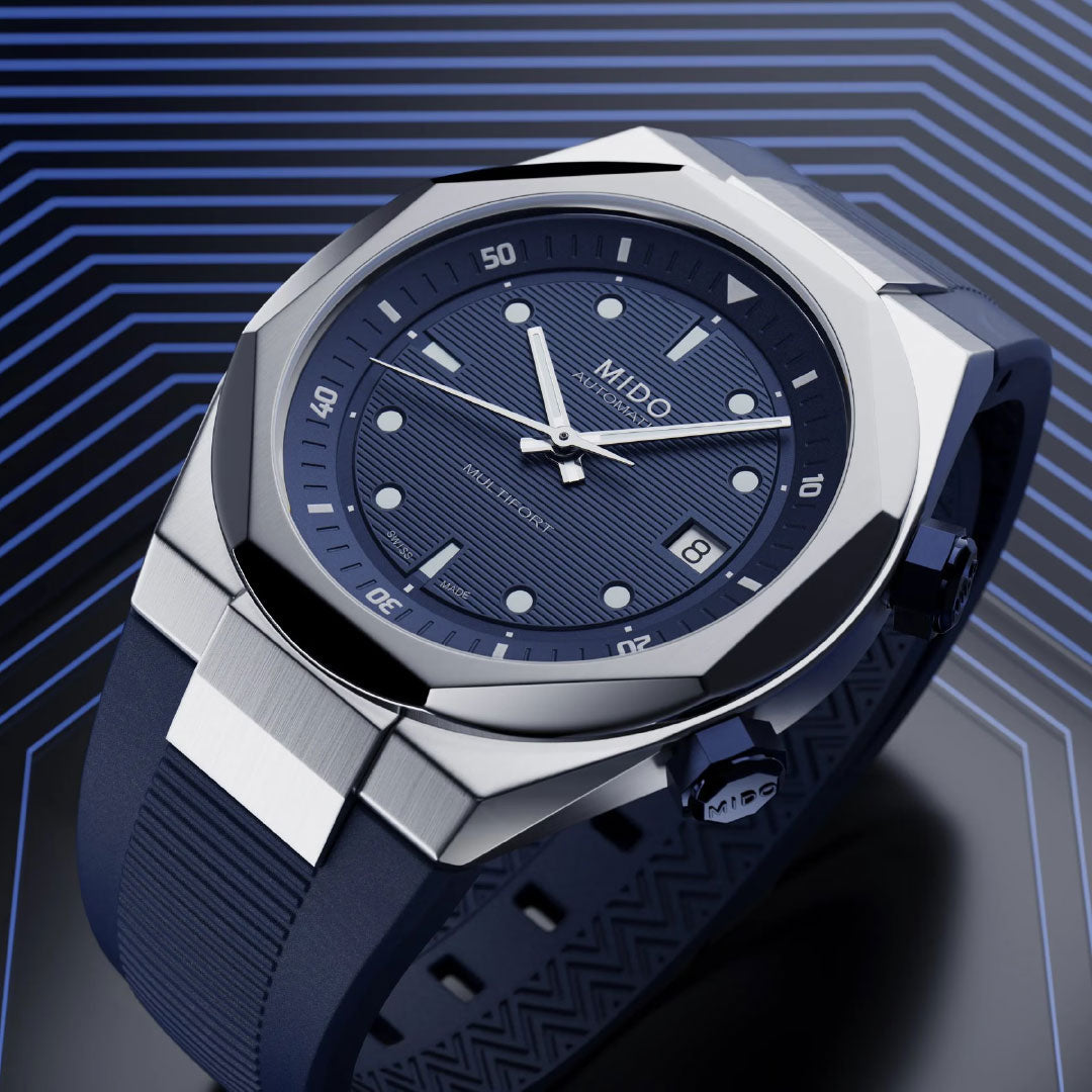 A close-up of the MIDO Multifort 8 Two Crowns Automatic 40mm watch, featuring a blue textured dial, silver hour markers, and a matching blue strap by MIDO, set against a geometric blue-toned background.