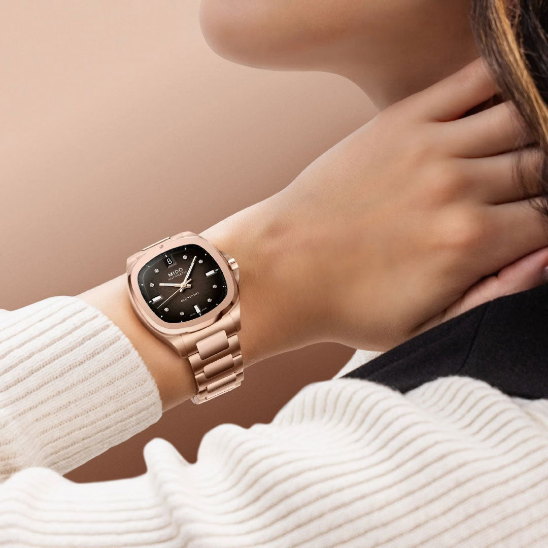 A person wearing the MIDO Multifort TV 35 Automatic 35mm watch with a rose gold case and black dial with silver markers touches their neck, dressed in a white ribbed sweater and dark jacket against a neutral background.