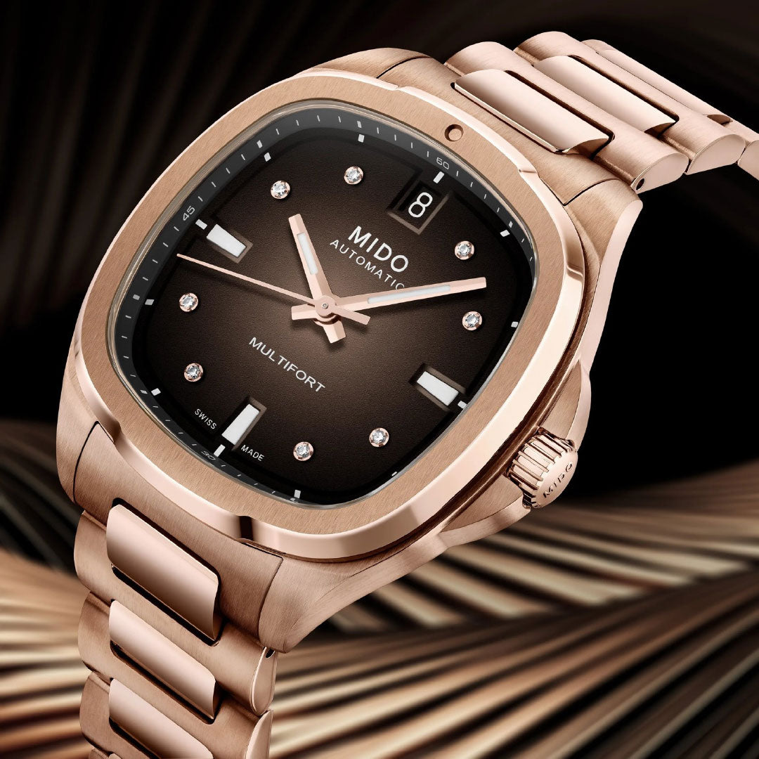 The MIDO Multifort TV 35 Automatic 35mm Watch in rose gold features a brown dial, diamond hour markers, date window at 3 o'clock, and a Nivachron balance spring, shown against an elegant blurred background.