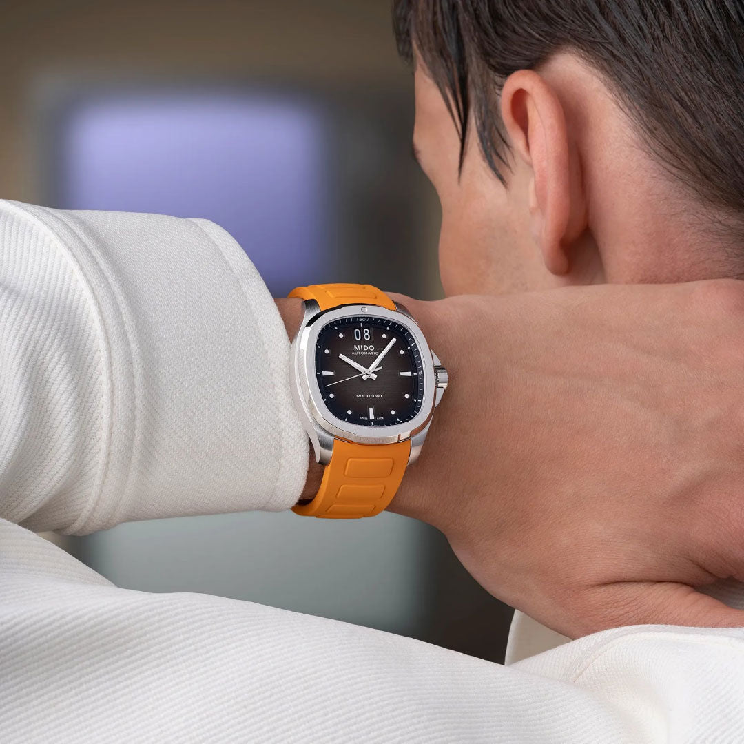 A person with short dark hair, seen from behind in a white shirt and an orange-banded MIDO Multifort TV Big Date Automatic 40mm Watch, holds their neck. The Swiss timepiece features a black dial with silver details.