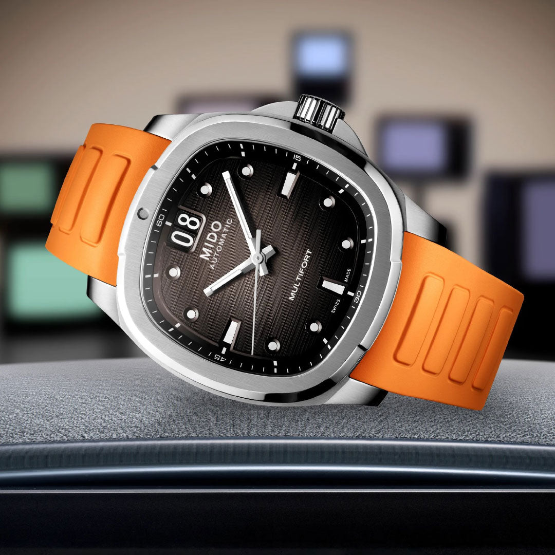 The MIDO Multifort TV Big Date Automatic 40mm Watch features an orange band, black textured dial, and silver case, highlighting Swiss craftsmanship with automatic movement and day-date display. It sits on a soft surface with blurred colorful rectangles behind.