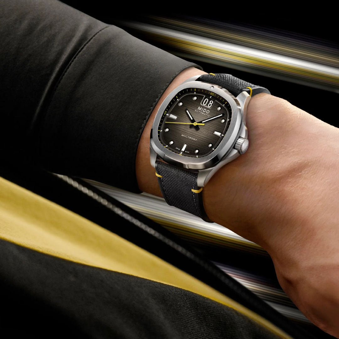 A close-up of a wrist wearing the MIDO Multifort TV Big Date Automatic 40mm Watch: black and silver case, yellow accents, textured dial, and black strap—a perfect match for their stylish black and yellow outfit.
