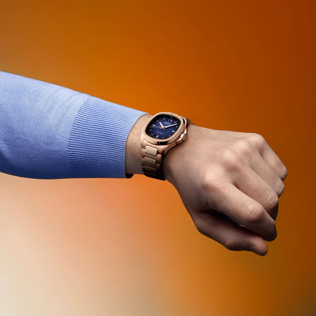 A person in a light blue long-sleeve shirt showcases the MIDO Multifort TV Big Date Automatic 39.2mm Watch, featuring a gold case and dark blue dial, set against a gradient orange background.