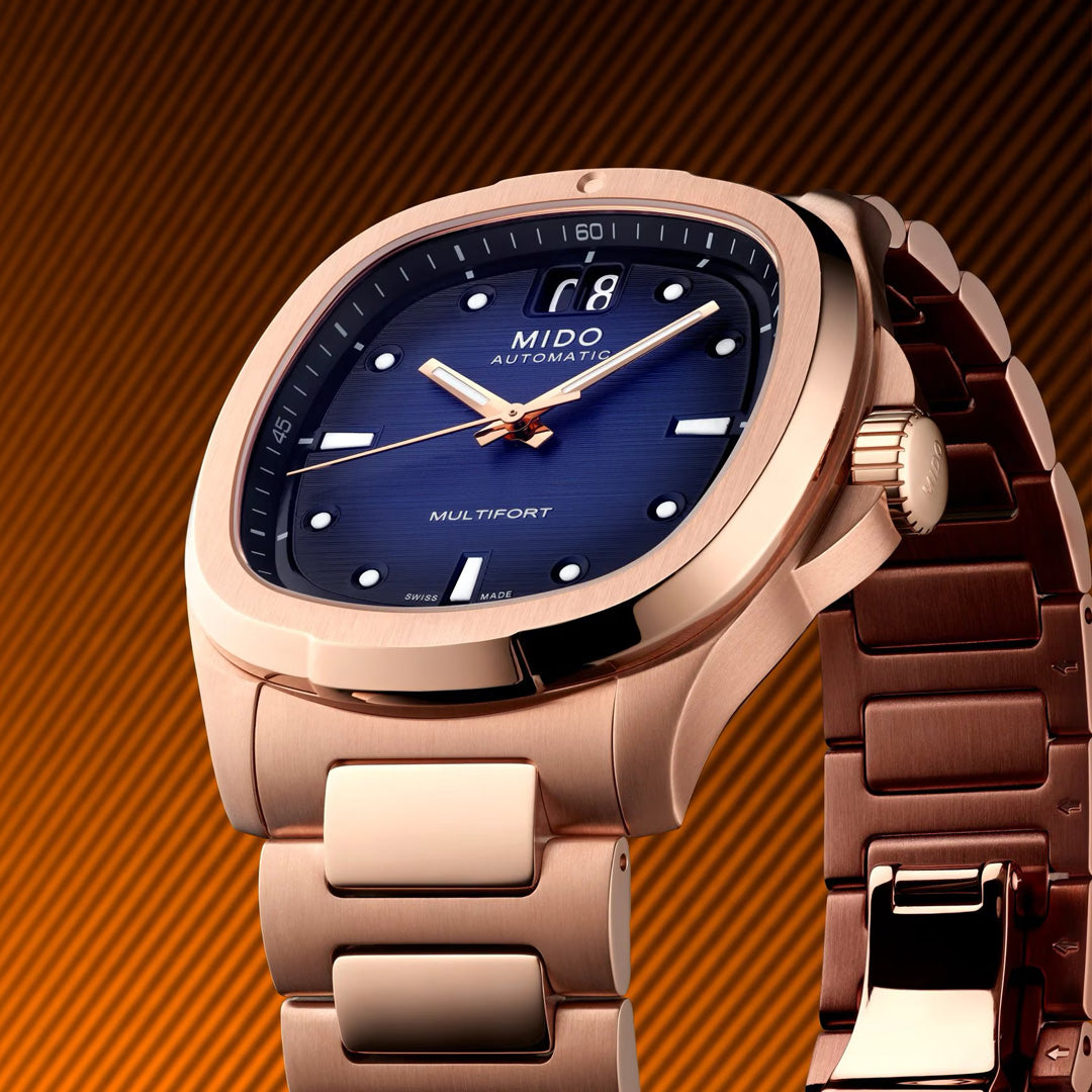 Close-up of the MIDO Multifort TV Big Date Automatic 39.2mm Watch, featuring a rose gold metal band, blue dial, date display, and luminous markers/hands by MIDO, set against an orange striped background.