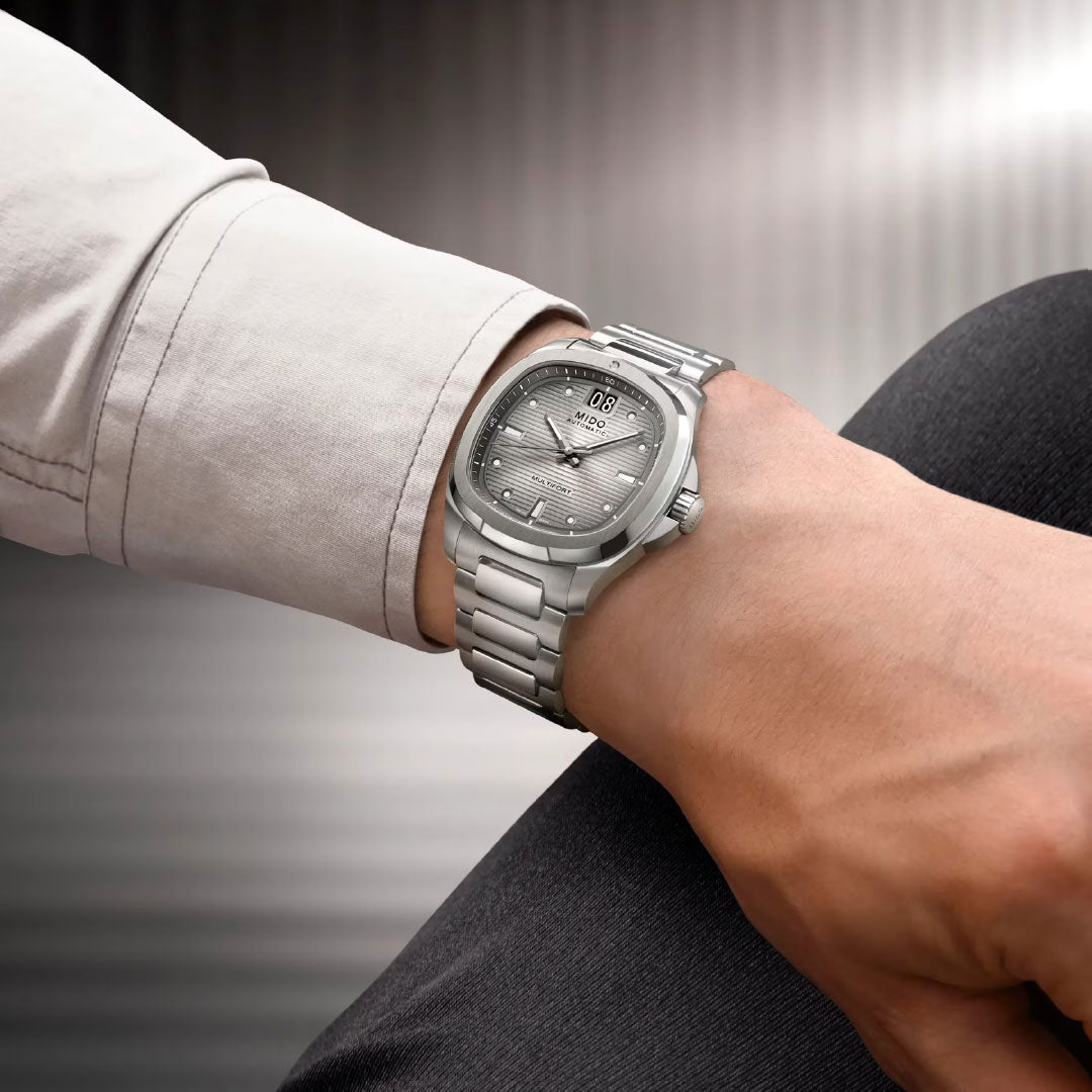 A person wears a MIDO Multifort TV Big Date Automatic 40mm Watch with a metal band and textured dial on their left wrist, dressed in a light shirt and dark pants, set against a blurred striped background.