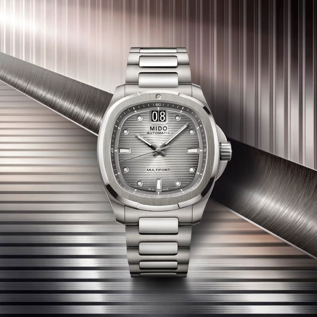 The MIDO Multifort TV Big Date Automatic 40mm Watch features a metallic bracelet, grey dial with date display, and "Multifort" branding. Displayed against a striped background, it highlights the distinctive design of MIDO.
