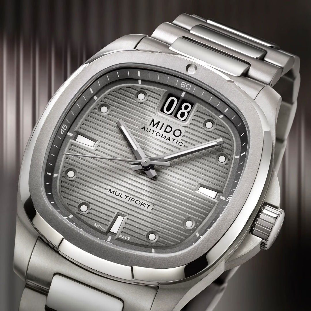 Close-up of the MIDO Multifort TV Big Date Automatic 40mm Watch, featuring a textured grey dial, bold markers, date at 12 o’clock, automatic movement, and metallic strap by MIDO, set against a blurred striped background.