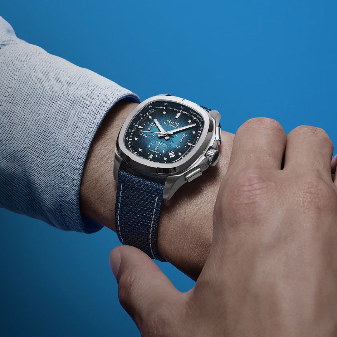 Close-up of a wrist wearing the MIDO Multifort TV Chronograph Automatic 42mm Watch with a blue face and textured strap, paired with a light blue shirt against a solid blue background.