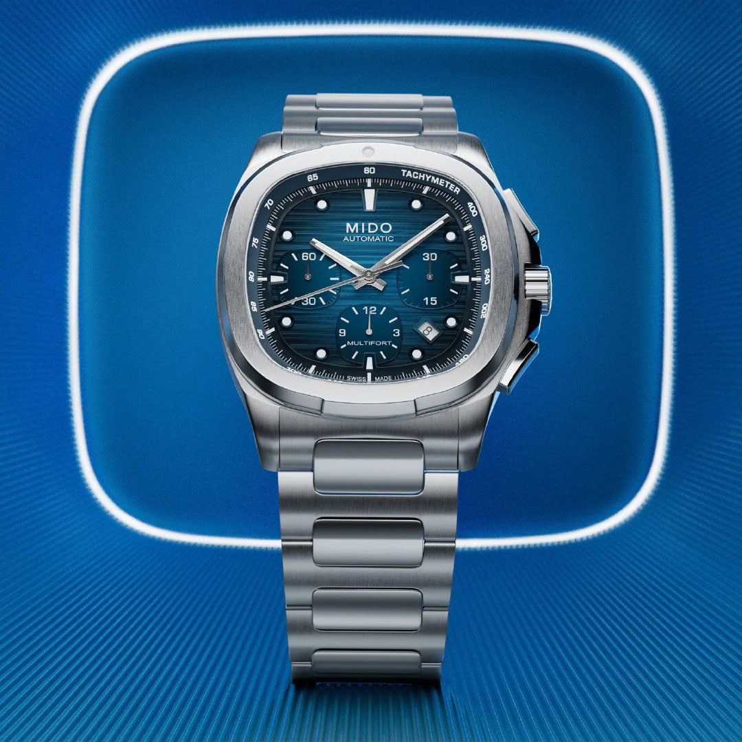 The MIDO Multifort TV Chronograph Automatic 42mm Watch in silver, featuring a blue dial with chronograph functions, is displayed against a glowing blue square background with a radial pattern.