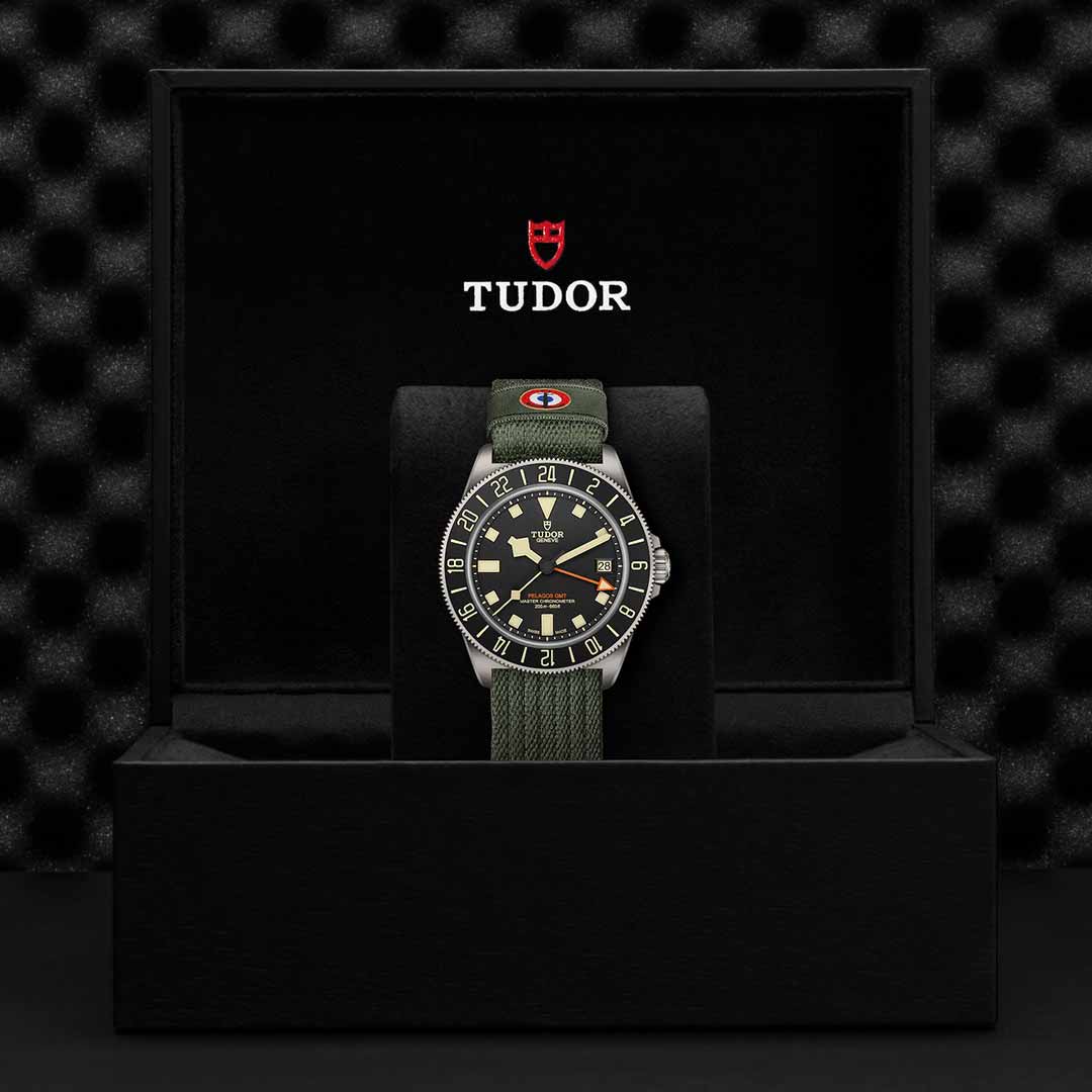 A TUDOR Pelagos FXD GMT watch with a green fabric strap is showcased in an open black box, exemplifying the precision of Zulu time. The watch face presents a black dial and tan hour markers, staying true to the Pelagos FXD GMT design. The TUDOR logo is prominently visible on the interior lid of the box.
