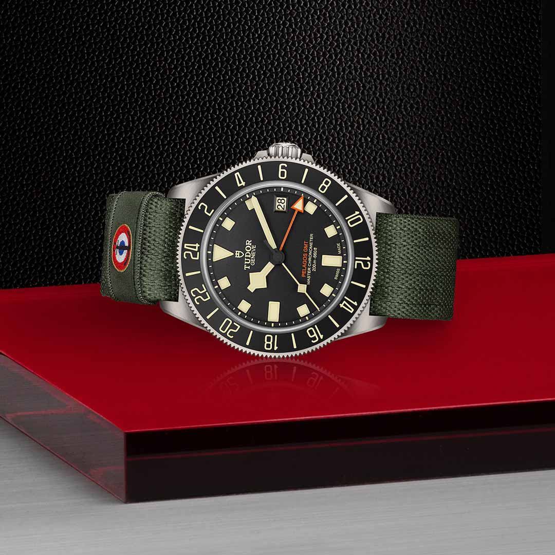 A TUDOR Pelagos FXD GMT watch, equipped with a green strap and black dial, is showcased on a red surface against a dark textured backdrop. This timepiece boasts bold hour markers, a rotating bezel, and military precision drawing inspiration from French Naval Aviation, making it ideal for tracking Zulu time.