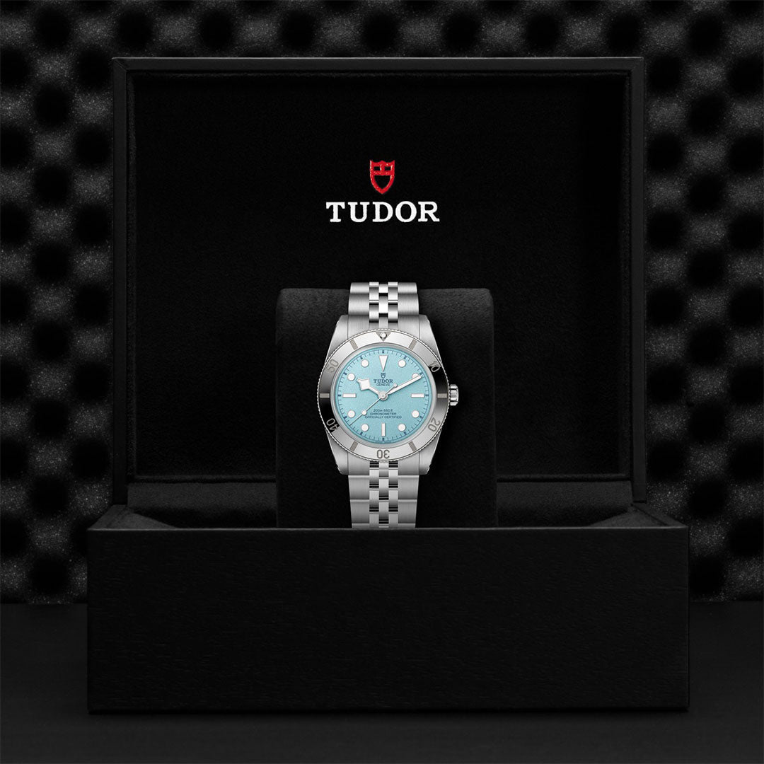 A TUDOR Black Bay 54 Lagoon Blue dive watch with a light blue dial is upright in a black presentation box, displaying the white TUDOR logo and red shield on the inside lid. The textured background is black.