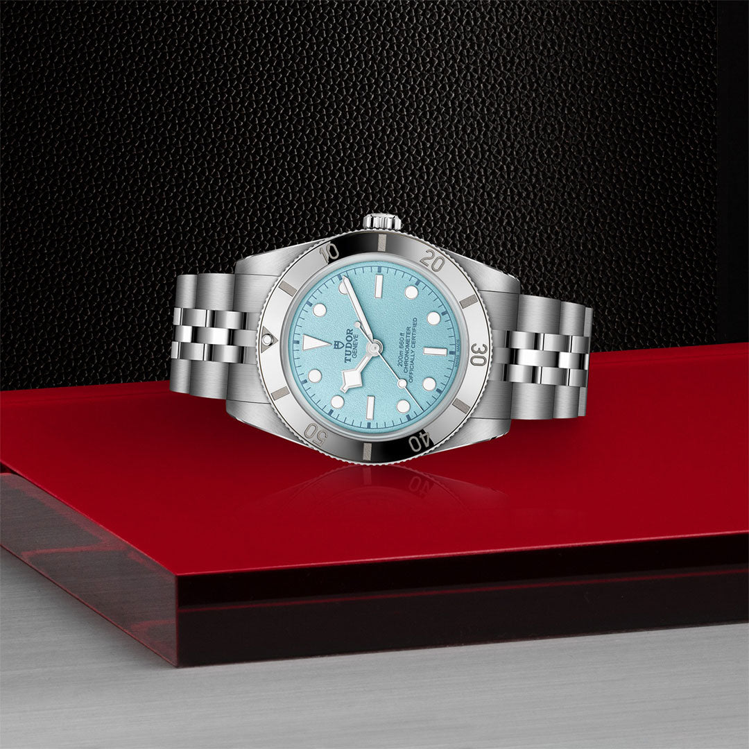 A TUDOR Black Bay 54 Lagoon Blue dive watch with a silver metal link band and light blue face rests on a glossy red and gray geometric surface, set against a textured black background.