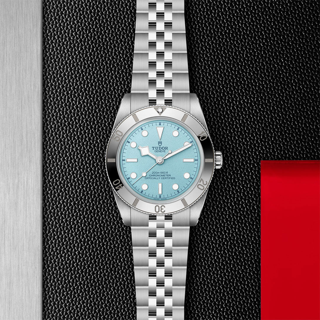 The TUDOR Black Bay 54 Lagoon Blue, featuring a light blue dial and metal bracelet, is displayed upright on a textured black surface with metallic and red accents.