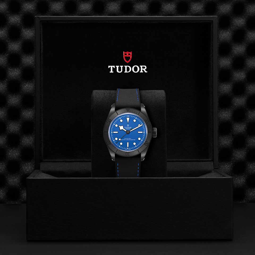 A TUDOR Black Bay Ceramic watch, showcasing a blue dial and black strap, is placed in an open black box. The box, adorned with the TUDOR logo and name in white and red, sits on a textured black surface, emphasizing the watch's Manufacture Calibre precision.