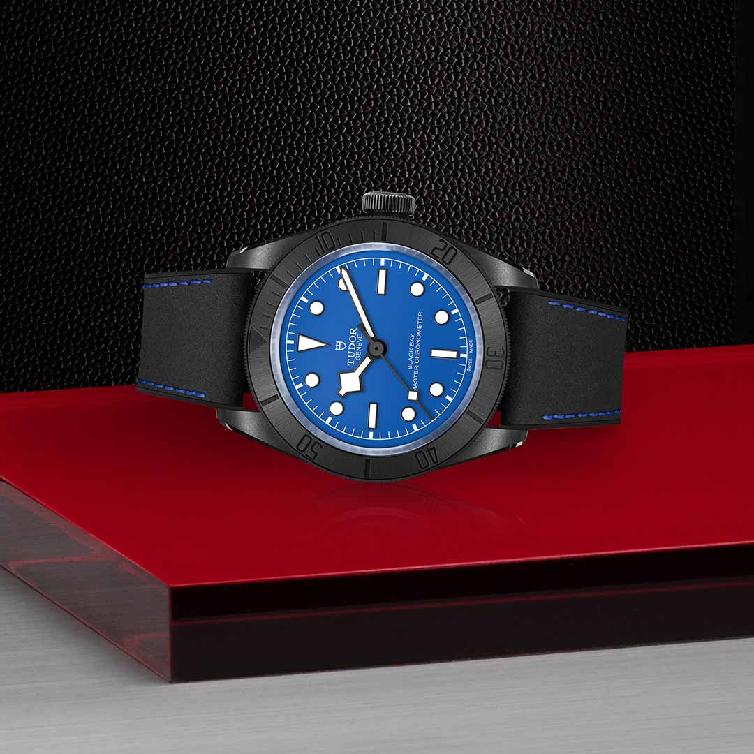 The TUDOR Black Bay Ceramic, featuring a striking blue face and a sleek black rubber strap with a ceramic case, is elegantly showcased on a glossy red and black surface. Its modern sophistication is further enhanced by the textured black wall backdrop, perfectly capturing the precision synonymous with TUDOR's Master Chronometers.