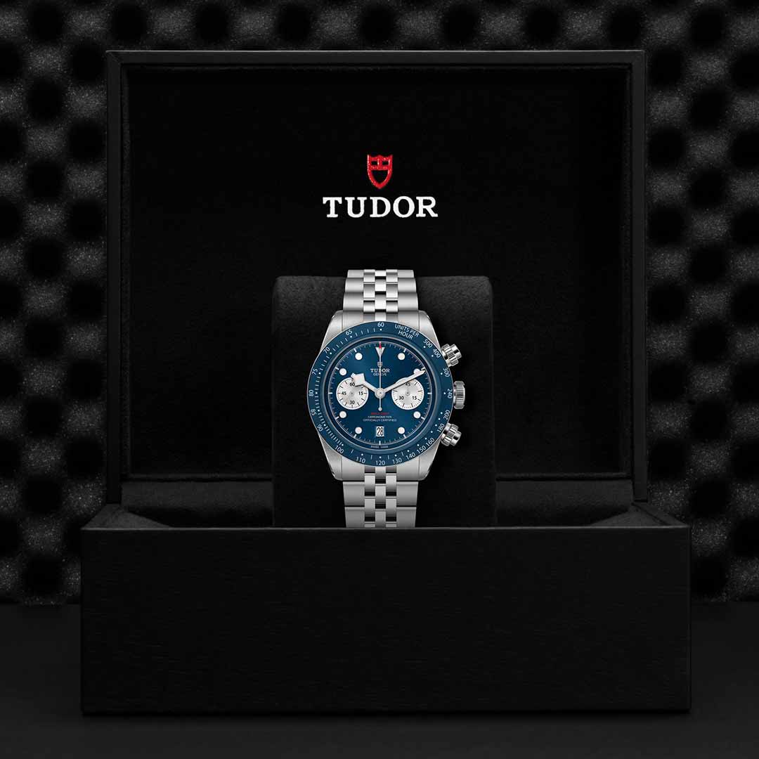 A TUDOR Black Bay Chrono Blue watch with a blue dial and silver subdials is displayed upright in an open black Tudor box, featuring the TUDOR logo and name on the inside lid.