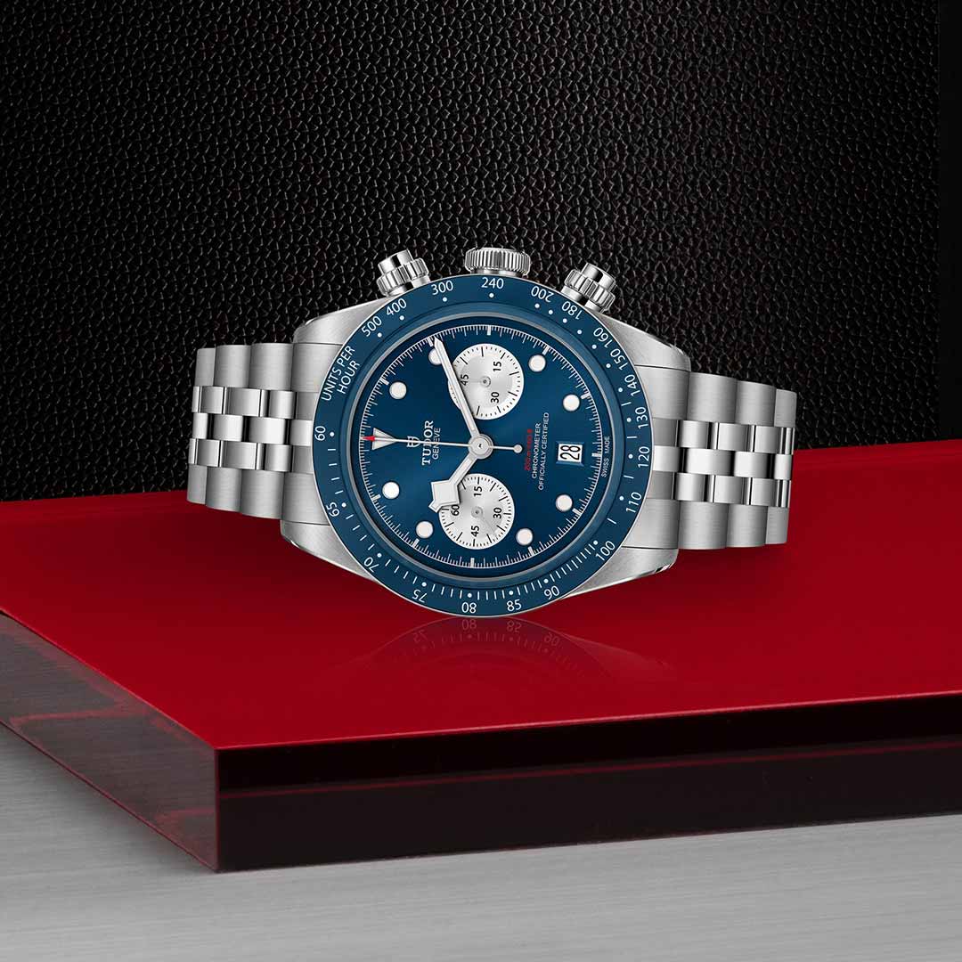The TUDOR Black Bay Chrono Blue, a boutique-exclusive, features a stainless steel case, blue dial with two white subdials, and is displayed on a red and silver surface set against a textured black background.