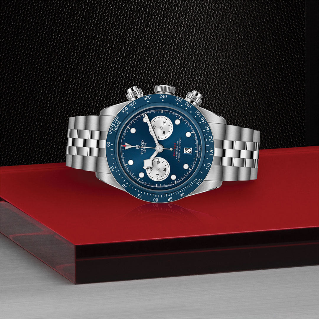 The TUDOR Black Bay Chrono Blue, a boutique-exclusive stainless steel luxury watch by TUDOR, features a blue dial with silver subdials, date window, and metal bracelet. It’s displayed on a red platform against a textured dark background.