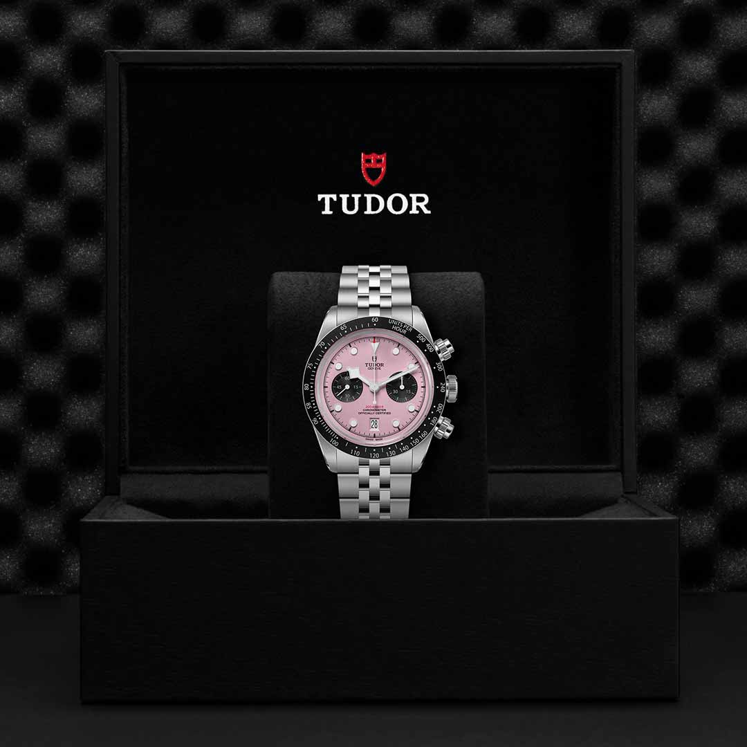 A TUDOR Black Bay Chrono wristwatch with a silver case, pink dial, and black subdials is showcased in an open black TUDOR box against a textured black background.