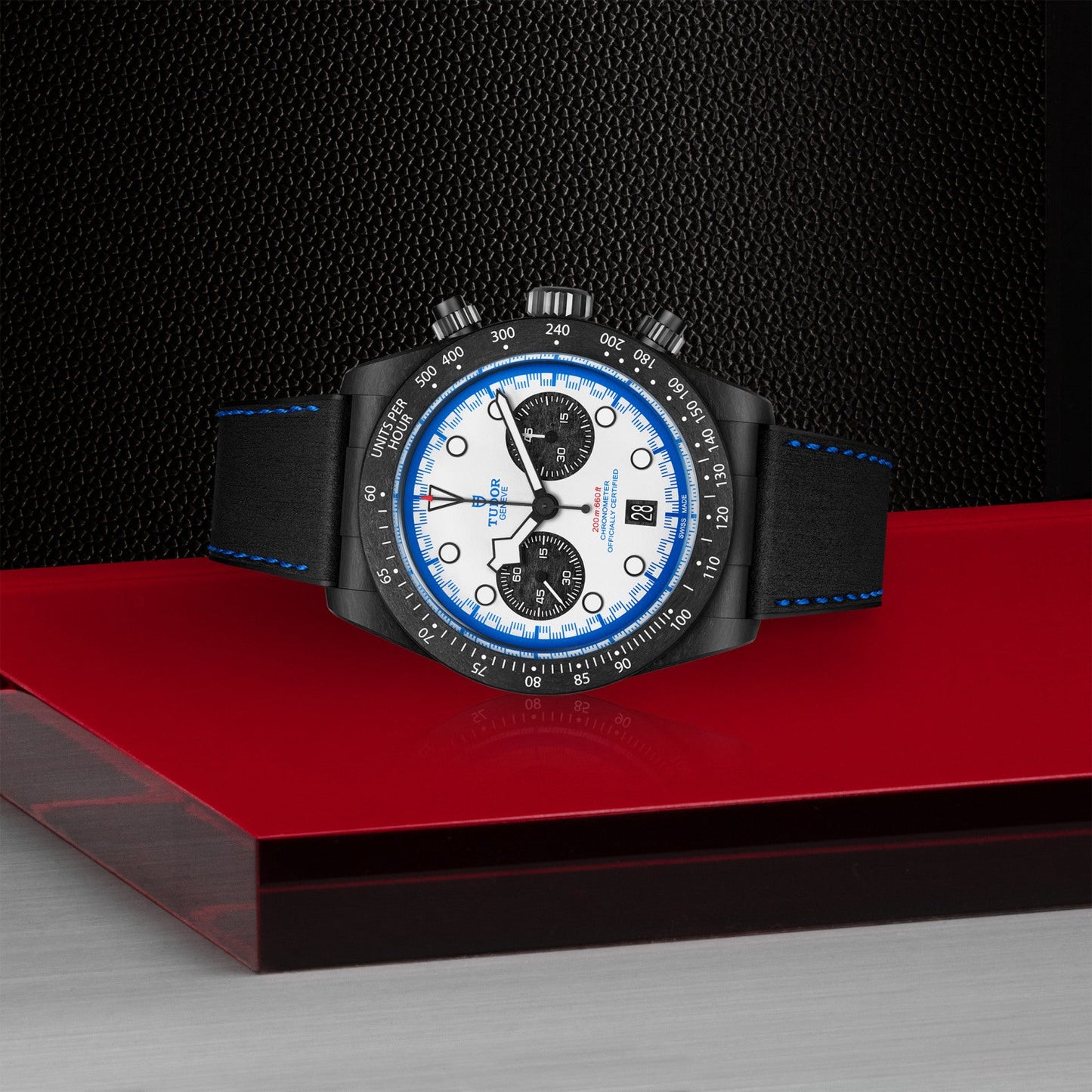 The TUDOR Black Bay Chrono “Carbon 25” wristwatch, with distinctive TUDOR Blue accents and a black leather strap, sits on a glossy red surface against a textured black backdrop for a modern, stylish look.