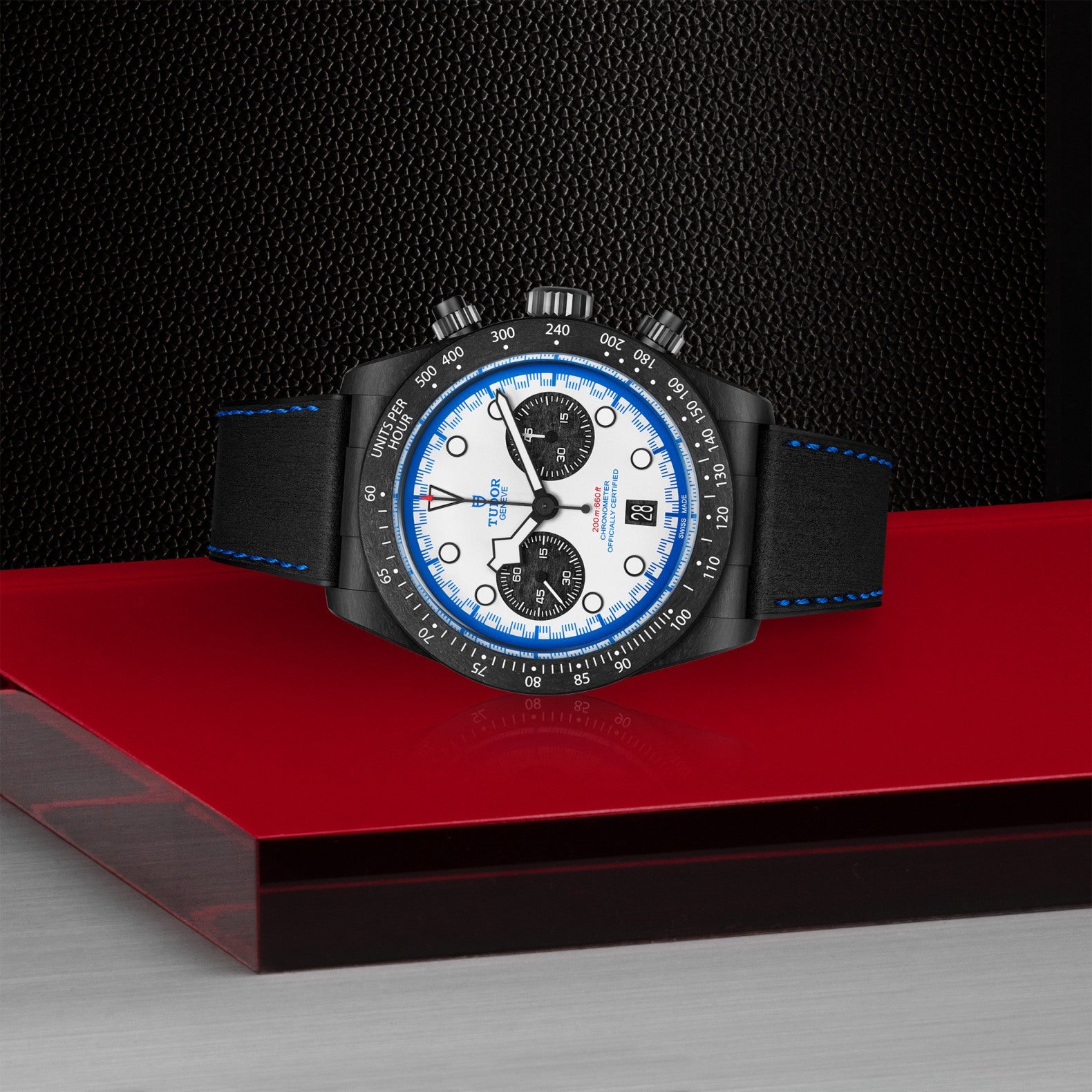 The TUDOR Black Bay Chrono “Carbon 25” wristwatch, with distinctive TUDOR Blue accents and a black leather strap, sits on a glossy red surface against a textured black backdrop for a modern, stylish look.