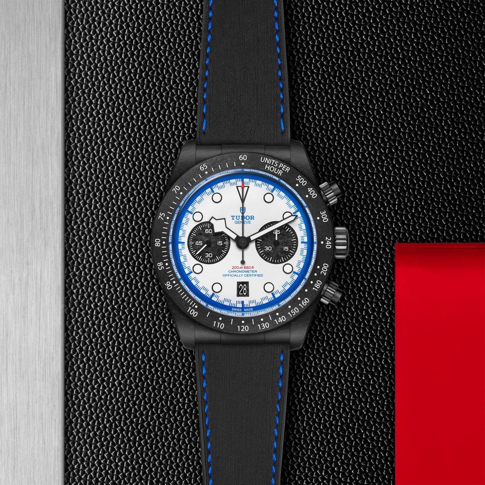 The TUDOR Black Bay Chrono “Carbon 25” features a black case, white dial with blue accents, and a black leather strap with blue stitching, shown on a dark textured background with metallic and red touches.