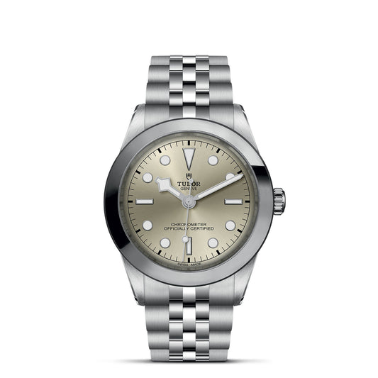 The TUDOR Black Bay One 39 features a 39mm steel case, metallic bracelet, and a light gray dial with minimalist black hour markers and hands. The brand name appears near 12 o'clock, and there's a date window at 3 o'clock.
