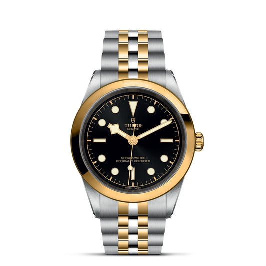 The TUDOR Black Bay One 41 S&G wristwatch features a round black dial with gold hour markers and hands. It has a 41mm steel case, with gold bezel and crown. The bracelet alternates silver and gold links, showcasing the "Tudor" brand name.