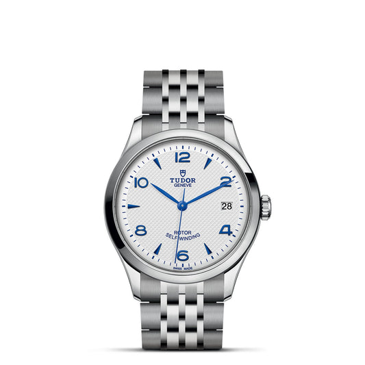 A silver timepiece from the TUDOR 1926 collection features a steel case, white textured face with blue Arabic numerals, and decorative blue hands. The dial displays "Tudor Geneve" and includes a date display, complemented by a stainless steel bracelet.