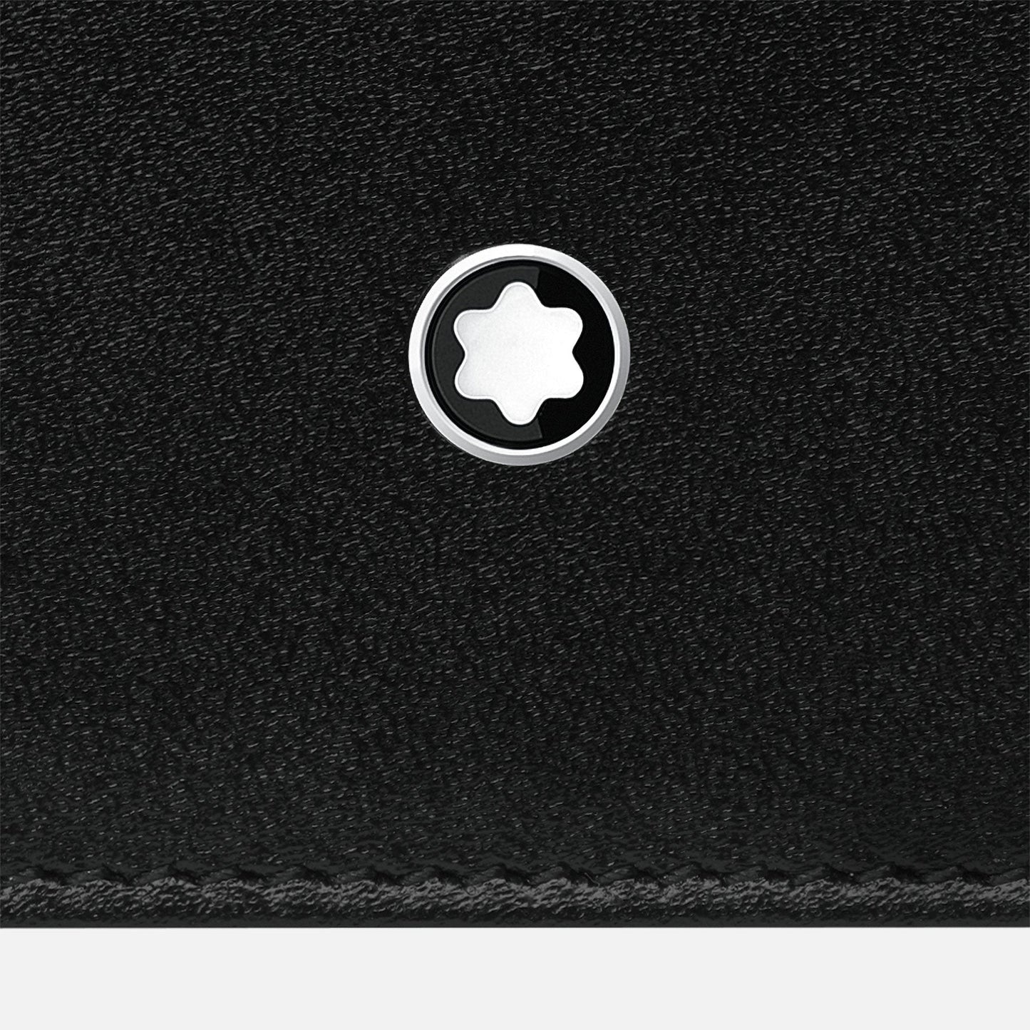 A close-up of black textured leather highlights an elegant accessory: a Montblanc Meisterstuck Pocket 6cc Leather Card Holder, featuring a compact, round silver emblem adorned with a white star-like symbol at its center. The lightweight emblem is positioned near the top edge above a line of stitching.