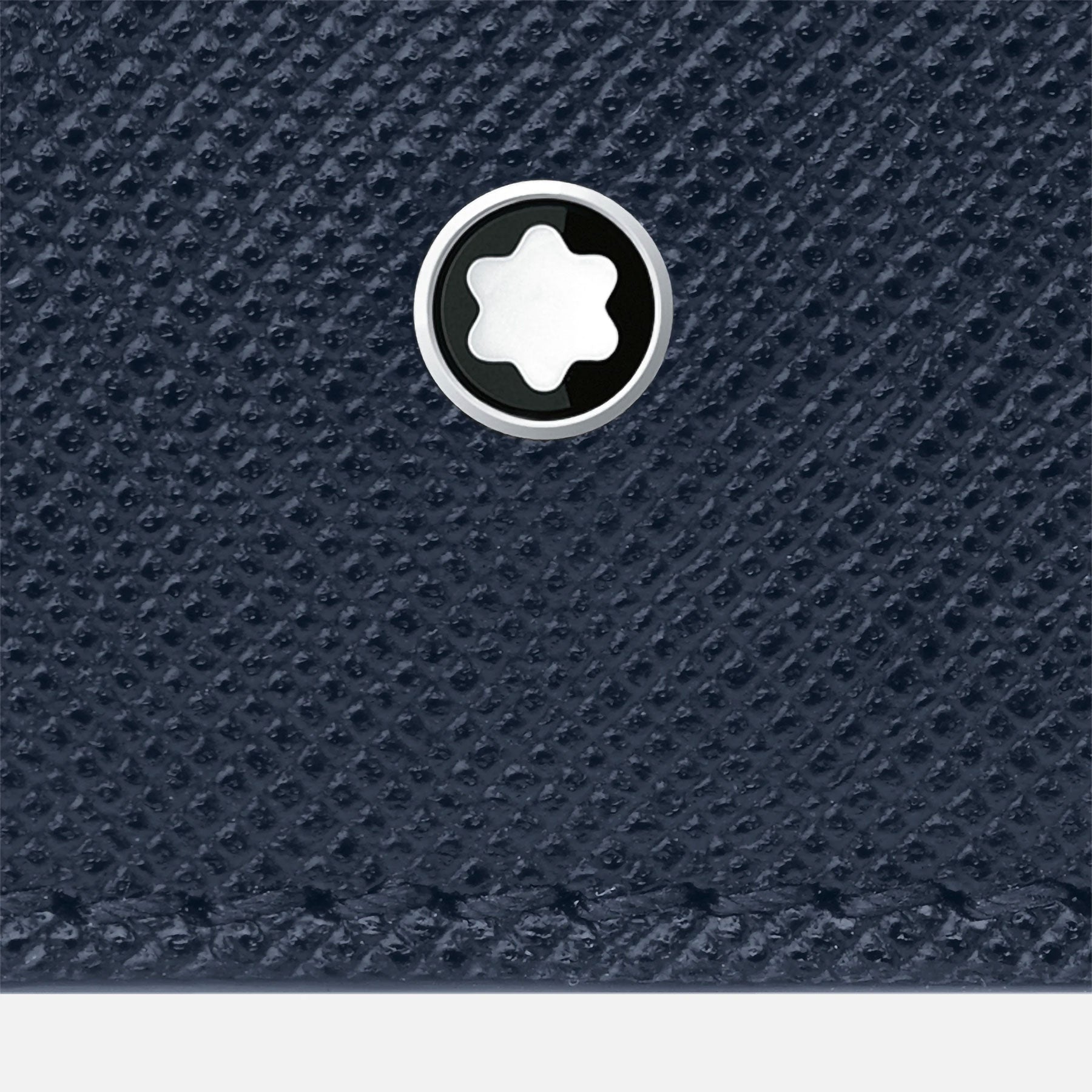 Close-up of a textured dark blue Saffiano leather surface on the Montblanc Sartorial Leather Wallet 6cc, adorned with a silver and black circular emblem with a star-shaped design in the center. This elegant piece is part of the distinguished Montblanc brand's Sartorial Collection.
