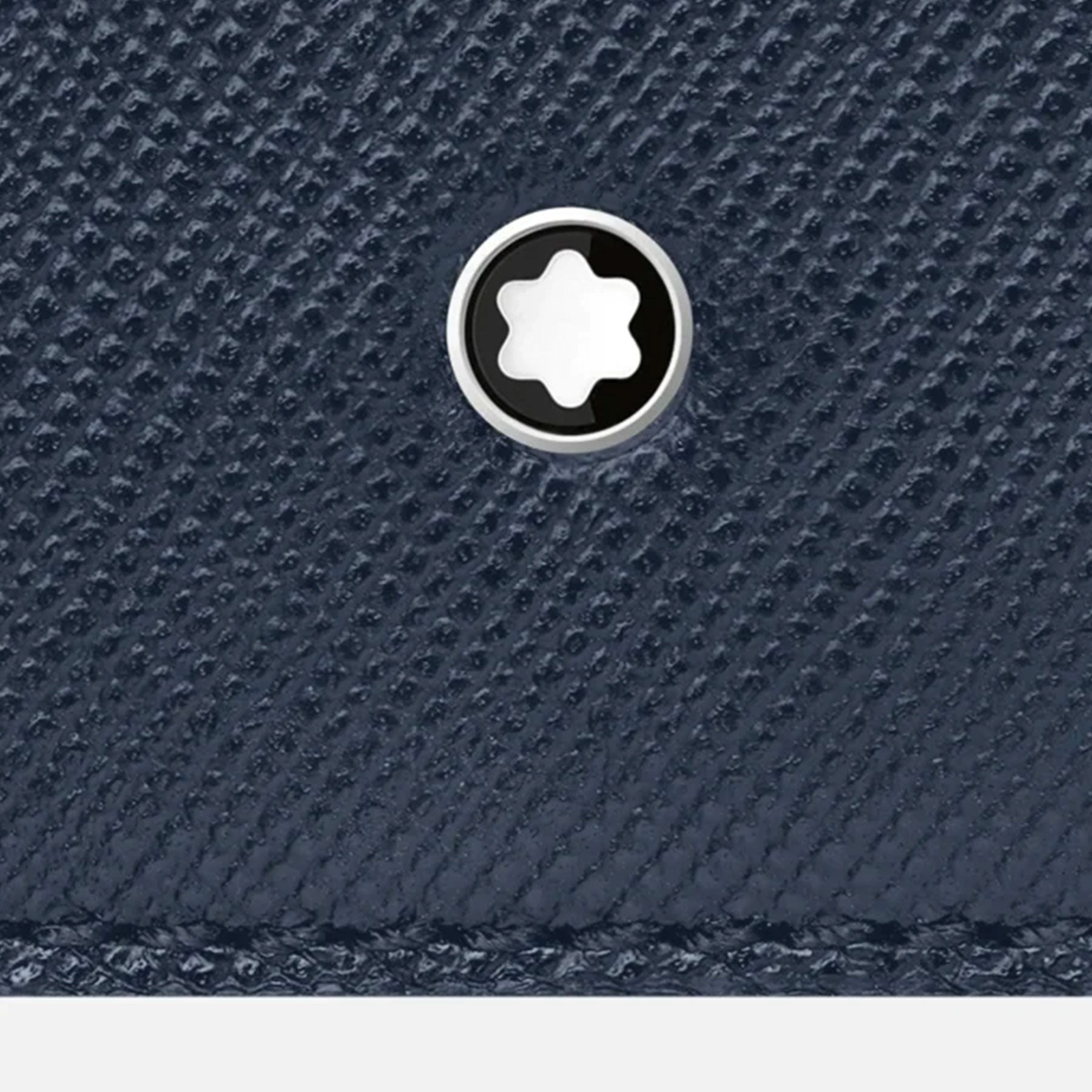Close-up of a textured dark blue leather surface, possibly from the Montblanc Sartorial Leather Business Card Holder, featuring a small circular metal emblem. The emblem displays a white star-like symbol on a black background, encircled by a silver border.