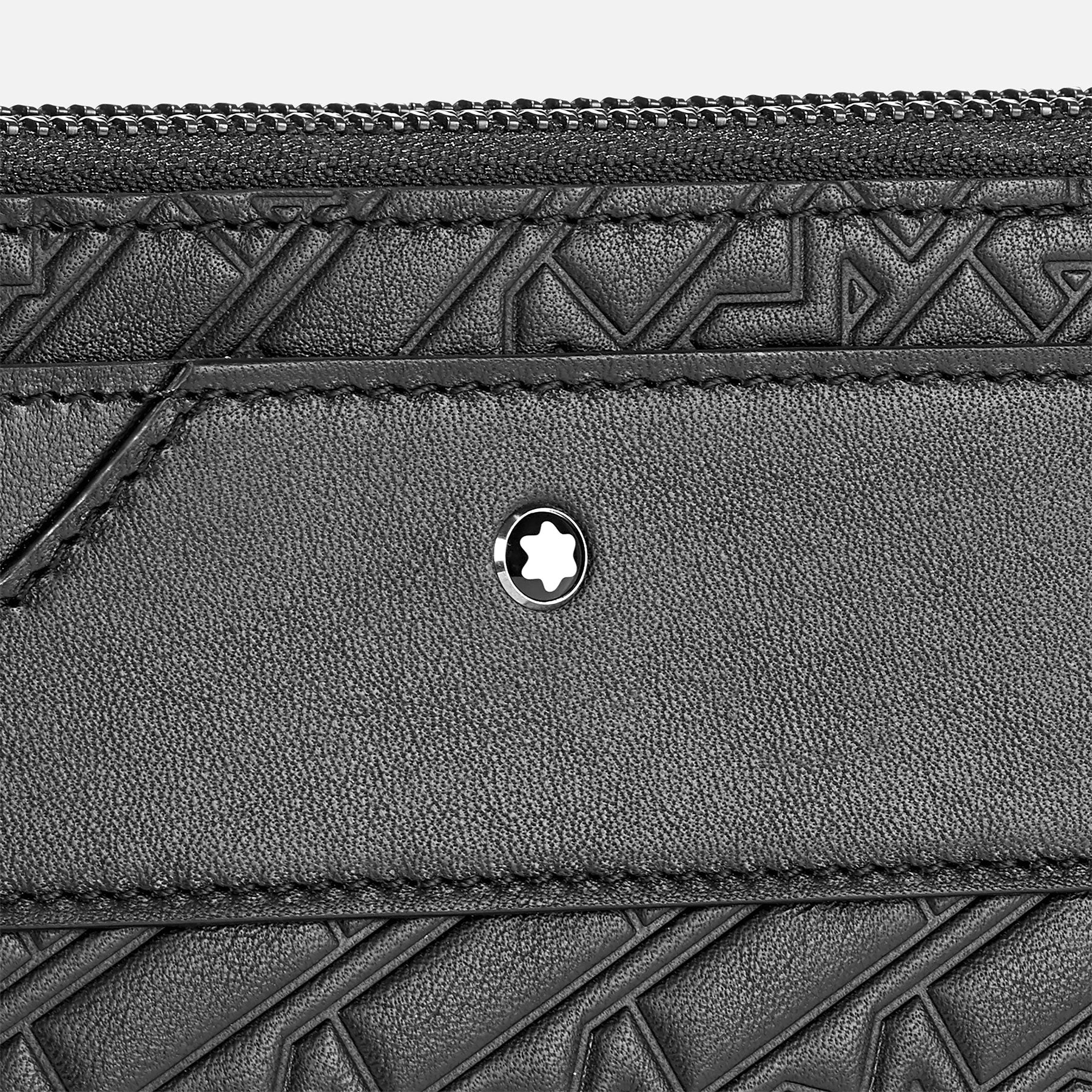 A close-up of the Montblanc M-Gram Leather Clutch Bag showcases its black textured leather with a geometric embossed pattern. Centered on the leather is a circular emblem featuring a star-like design. At the top, the stylish Montblanc M-Gram 4810 zipper is prominently visible.