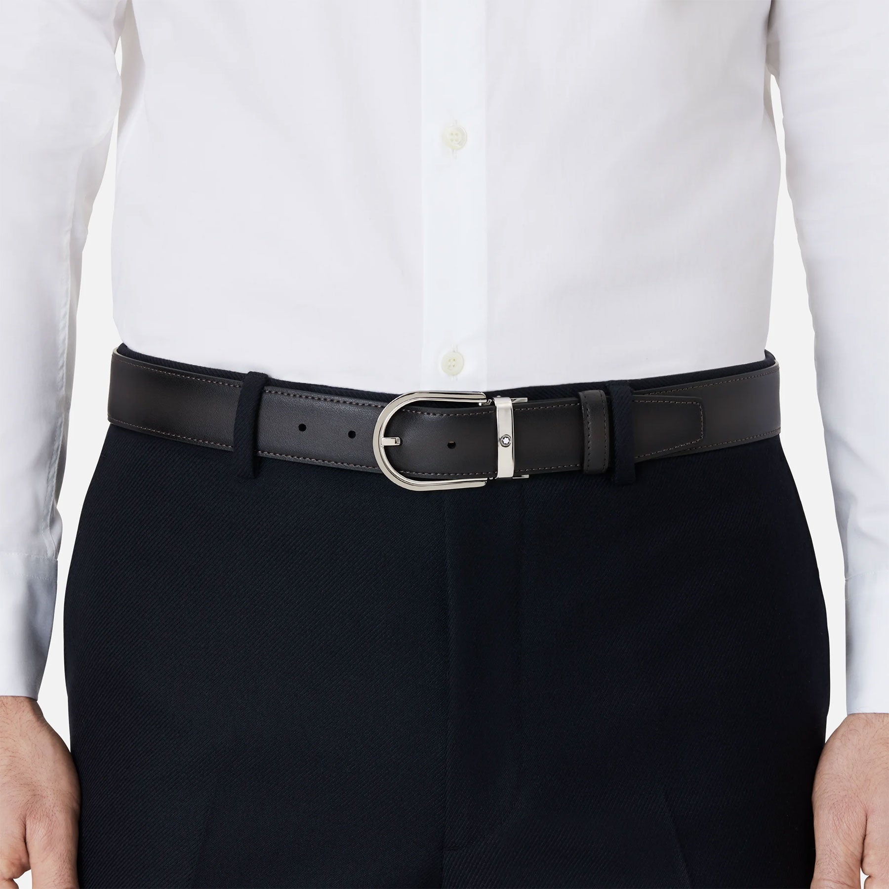A person wearing a white dress shirt and black formal pants, enhanced by the Montblanc Horseshoe Leather Belt, with its unique horseshoe buckle. The shirt is neatly tucked into the pants.