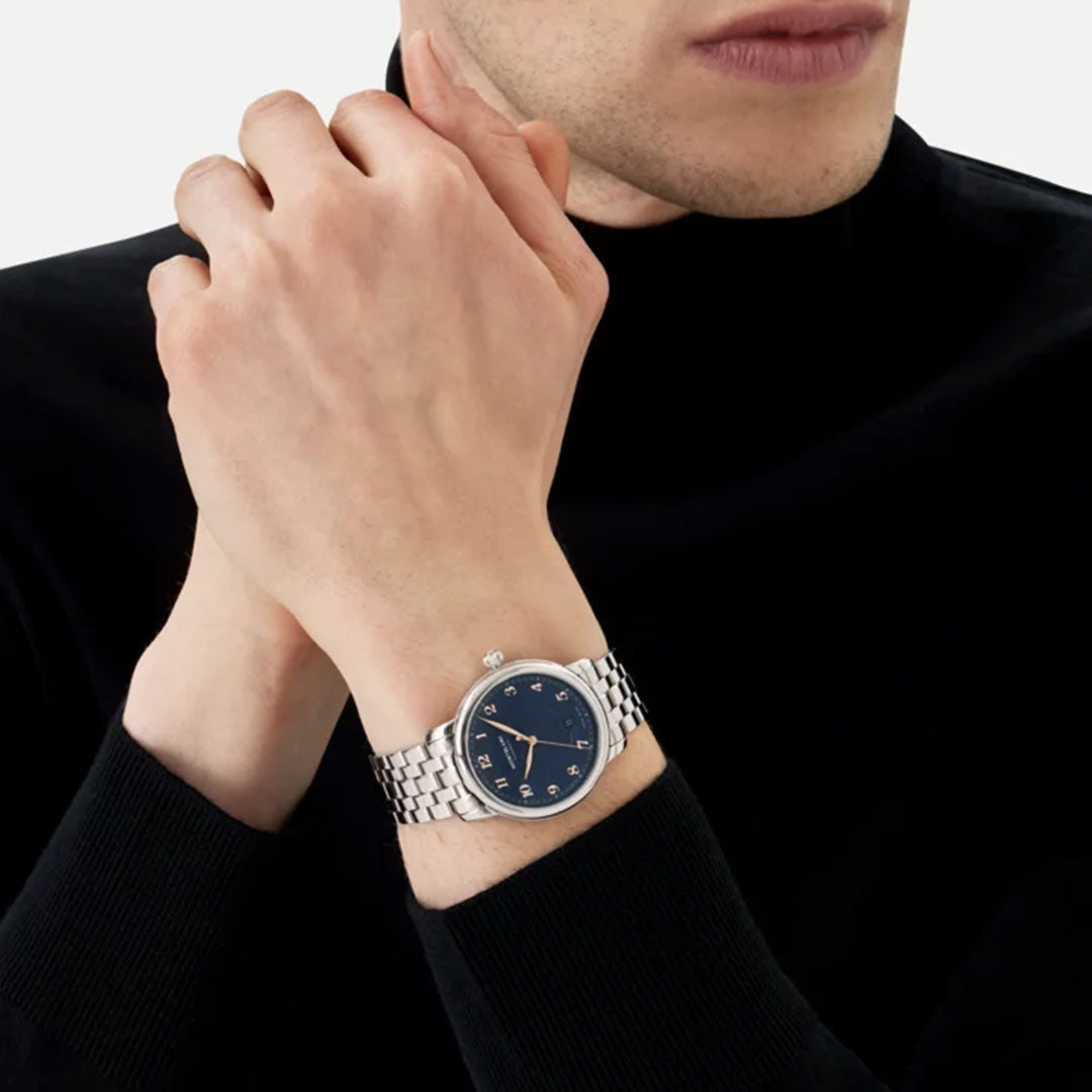 A person in a black turtleneck sweater with hands clasped, wearing a Montblanc Star Legacy Automatic 39mm Watch, which features a blue glacier pattern dial with Roman numerals. The light gray background enhances the elegance of the attire and timepiece.