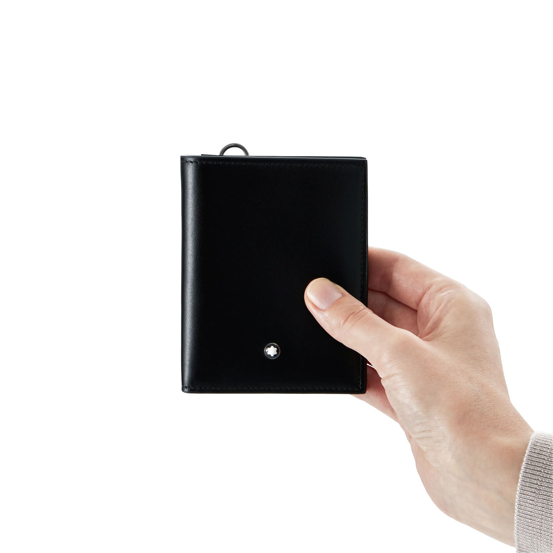 A hand holding the Montblanc Meisterstück Compact Black Leather Wallet 6cc against a white background. The closed wallet showcases a small metallic Montblanc logo on the front. The person is wearing a light-colored sleeve.