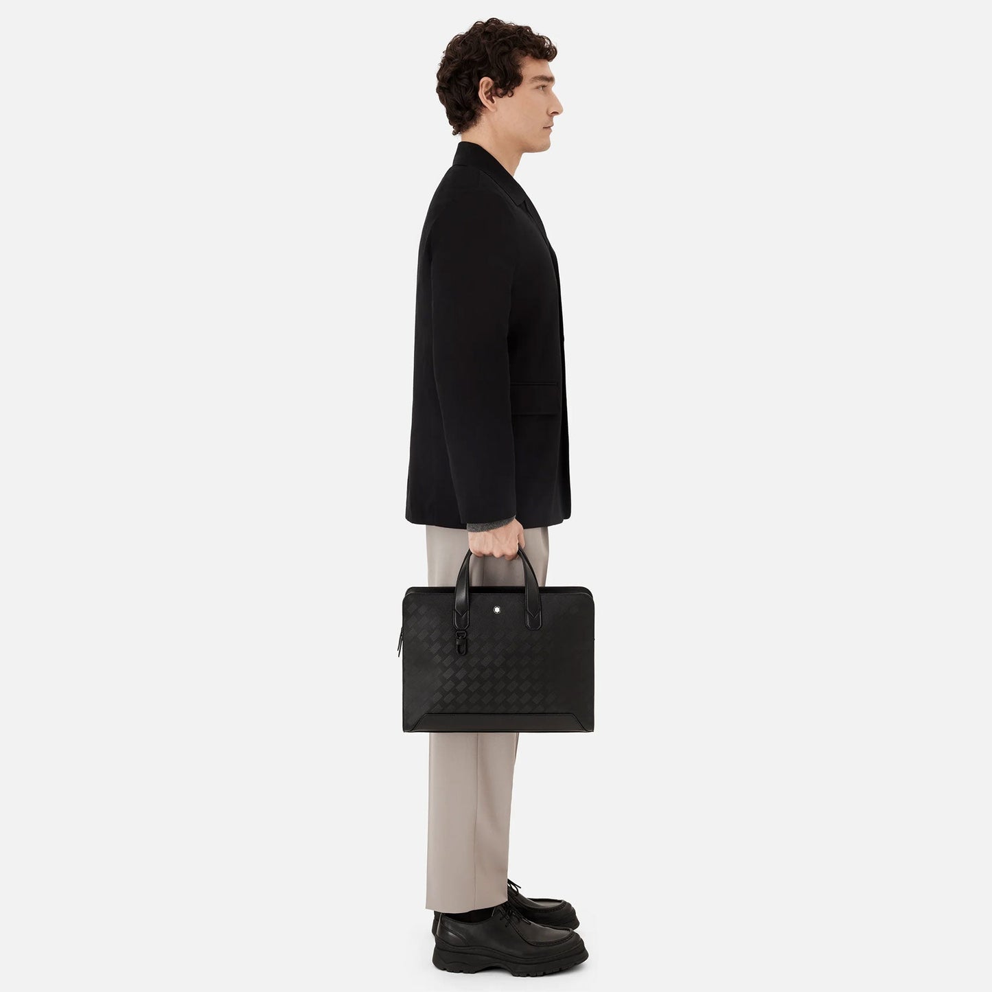The individual is posed in profile against a simple backdrop, dressed in a black jacket, beige pants, and black shoes. They are holding the elegant Montblanc Extreme 3.0 Black Leather Document Case by their sides with both hands.