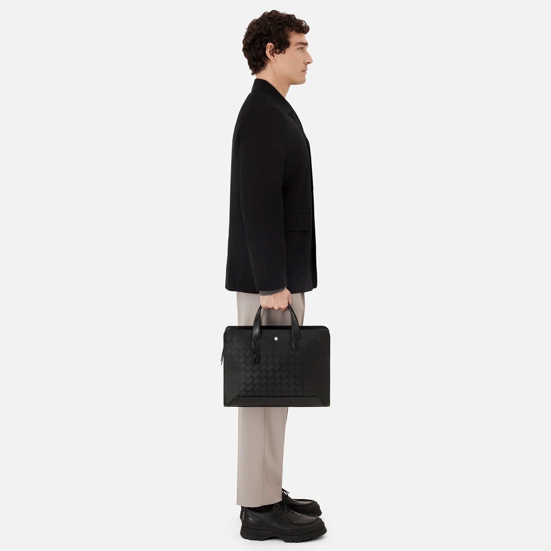 The individual is posed in profile against a simple backdrop, dressed in a black jacket, beige pants, and black shoes. They are holding the elegant Montblanc Extreme 3.0 Black Leather Document Case by their sides with both hands.